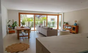 Salón moderno con comedor y sofá, destacando una pared prefabricada interior que se integra con grandes ventanales al jardín.
