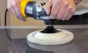 Manos puliendo una superficie oscura con una pulidora eléctrica. El disco de lana blanco gira para pulir madera.