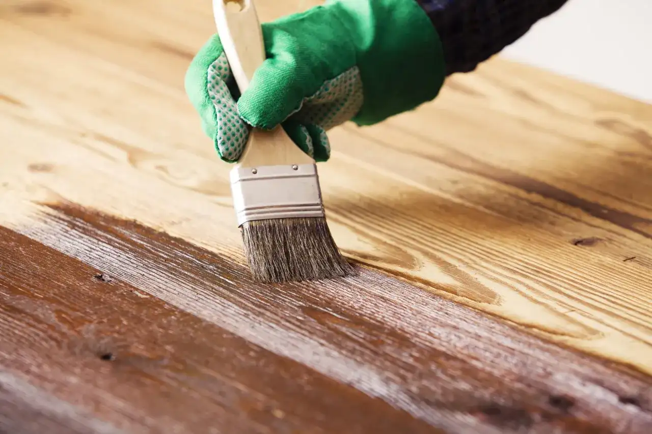 Mano con guante verde barnizando mesa de madera con brocha. El acabado protege y embellece la veta.