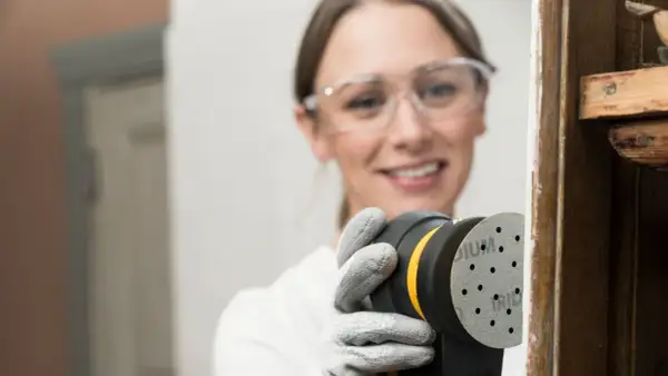 Mujer con gafas de seguridad usa lijadora orbital para restaurar madera, una forma eficiente de como lijar madera sin lija.